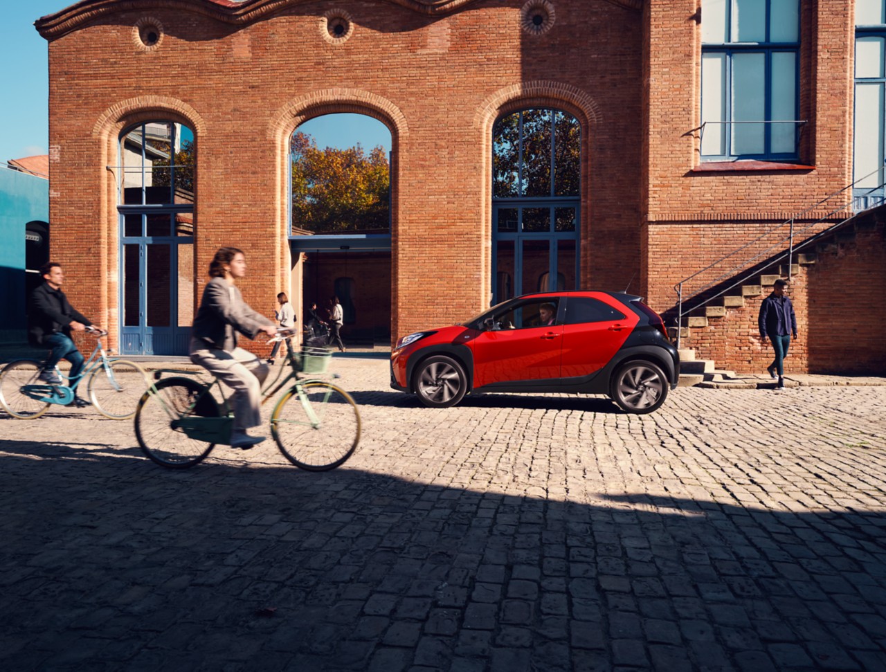 Image showing the side of Chili Red Aygo X parked at the side of the road and being passed by a cyclist from the left